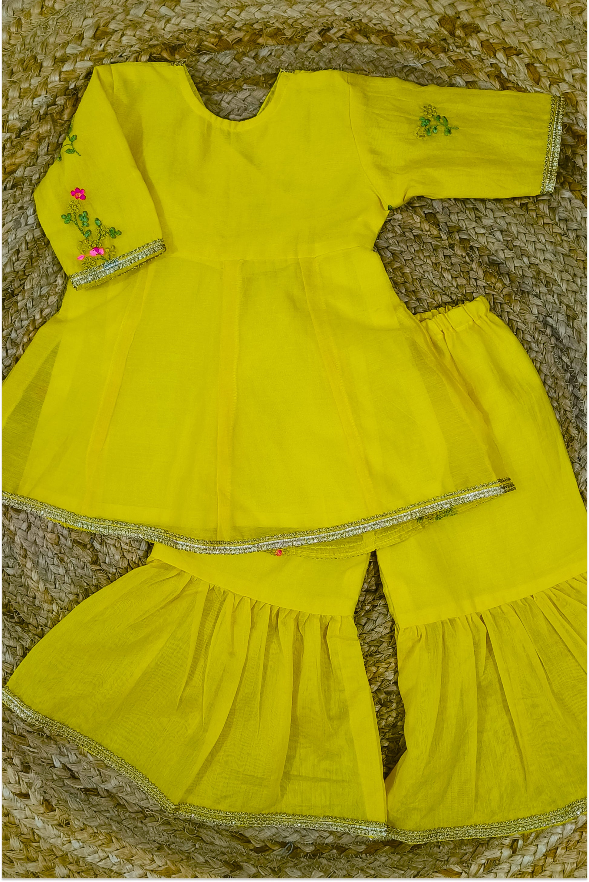Yellow mul chanderi sharara set flat lay showing peplum top with tie-up, sharara pants, hand embroidery and golden lace