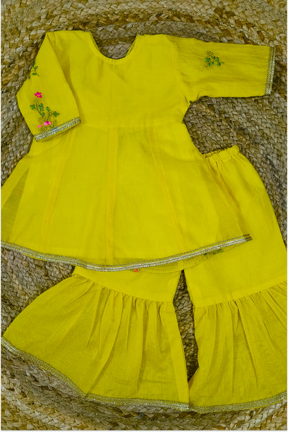 Yellow mul chanderi sharara set flat lay showing peplum top with tie-up, sharara pants, hand embroidery and golden lace